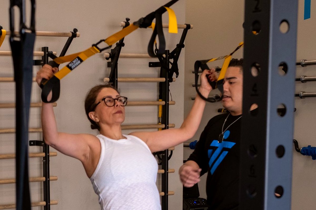 A woman in her forties mid-set on the suspension straps, an IMTI coach beside her watching the movement
