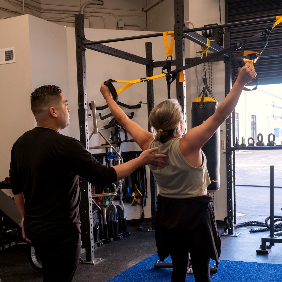 A coach cueing a client through an overhead press on the suspension trainer, a hand at her back