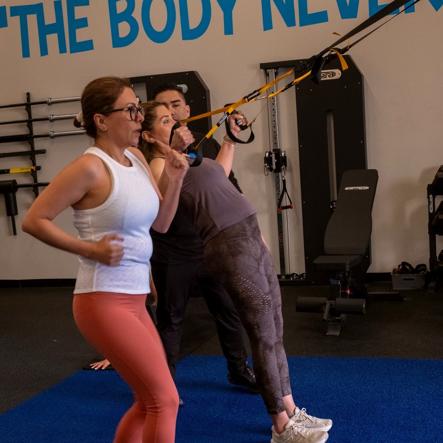 A client mid-stride at the step in the gym, a second client working the suspension trainer with a coach behind her