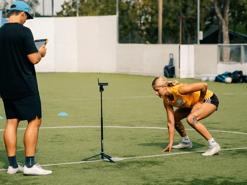 Athlete in a sprint-ready stance at a timing gate while a coach records the result