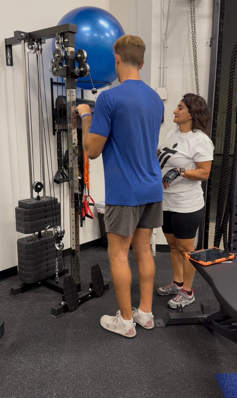 An IMTI coach at a member's side, watching him work the cable column, a tablet ready on the bench beside her