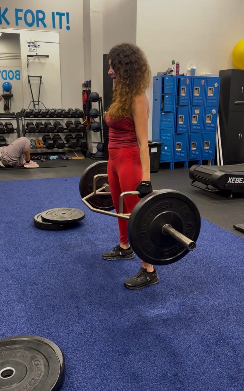 A member lifting a loaded trap bar on the turf while another member works on a mat behind her